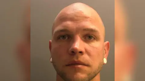 A mugshot of Sven Halstein. He has blue eyes, a shaved head and is wearing large white earrings covering both his lobes. 