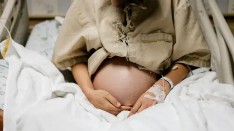 Close-up of belly a heavily pregnant person in a hospital bed, with an IV attached to her arm.