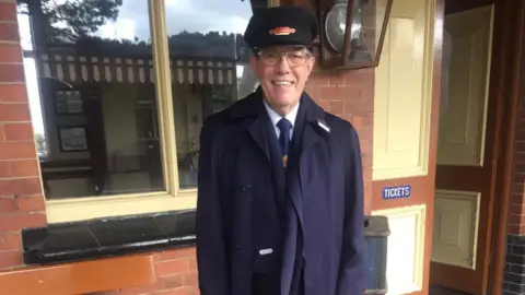 Clive Hughes Clive, dressed in a railway uniform, all in blue, with a hat on. He is standing by a railway station office, is smiling and there is an old fashioned lamp to the side of his head. A window and a doorway is behind him. 