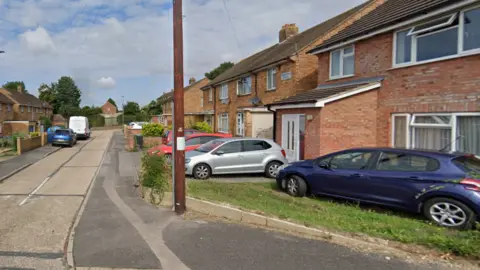 Google Terraces of brick houses and cars in Cinderford Close in a Google Street View image.