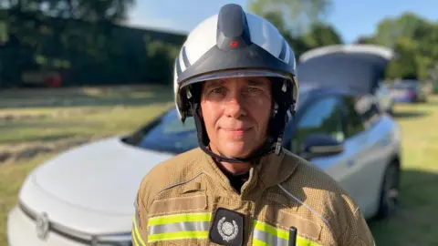 Luke Deal/BBC Gary Smart wears a large white helmet with a retracted visor, and brown firefighting jacket. He looks at the camera with a friendly stare and stands in front of a silver car on grass.