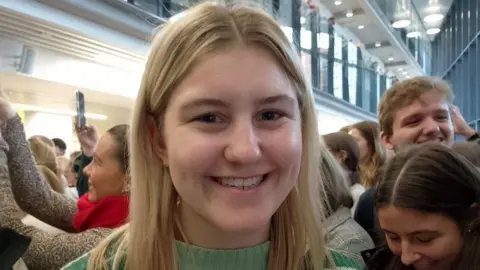 Leah Reid has long blond hair, is smiling and wearing a green jumper with other students standing behind her, some of them holding up their phones 