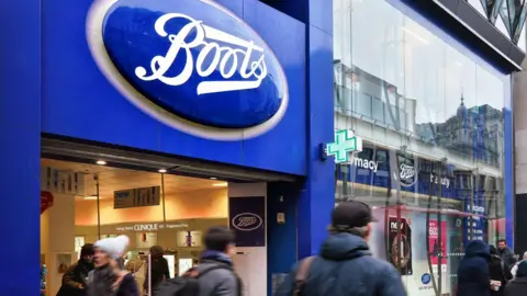 The entrance of a large Boots store with pedestrians walking in from of the large glass store. The blue Boots signage hangs above the door. 