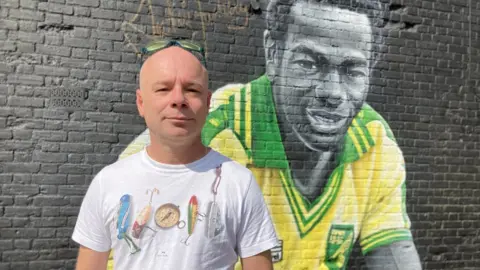 Andy Trigg/BBC The landlord of the Fat Cat and Canary pub stood in front of a Justin Fashanu mural