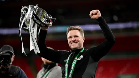 Eddie Howe holding the Carabao Cup aloft at Wembley. He has short fair hair and stubble and is wearing a black Newcastle United top. He is holding the silver trophy in his right hand. It is decorated with black-and-white ribbons.