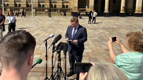 Council leader John Cotton reading a statement in front of microphones in front of Birmingham's Council House. 