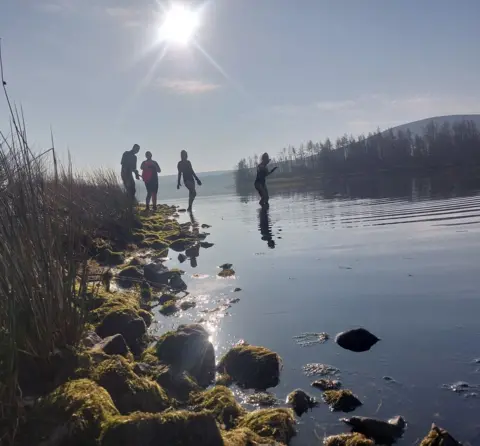 Raymond Carstairs Four wild swimmers stand in a reservoir, with stones lit by the sun in the foreground.