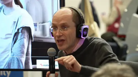 Tim Leunig, a bald middle-aged man with spectacles, holds a microphone and uses his finger to make a point during an impassioned speech at a Labour fringe meeting