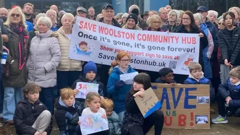 Large crowd of people at a protest with 10 children crouching in front on the ground holding some crayon-drawn signs saying "save the hub". The adults stand behind with a large banner saying "Save Woolston Community Hub - Once it's gone, it's gone forever".