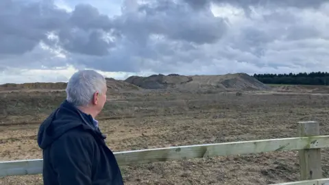 BBC Stephen Adkins standing next to a fence looking over his shoulder at a mound of mud in the distance