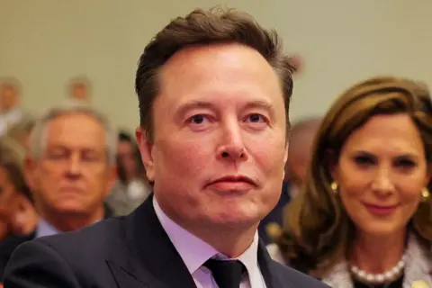 Reuters Elon Musk stares just above the camera, dressed in a a black suit, tie and white shirt. He has light brown hair and dark eyes, a crowd of people stand behind him. 