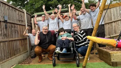 Oscar is in a wheelchair with a parent on either side. Behind them are a team of volunteers in grey t-shirts cheering.