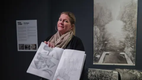 Juliette Losq, one of the artists' whose work is being displayed, is holding a sketchbook with a sketch inside, she is stood next to a larger piece of art on a canvas. She is wearing a scarf and her hair is in a ponytail