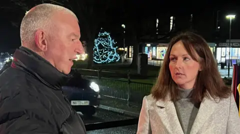 BBC Jim Cunningham and Lola McEvoy standing outside on the street, speaking to each other. There is a Christmas tree with blue lights in the background. It is dark and street lights are on. Mr Cunningham has grey hair and is wearing a thick black coat. McEvoy is wearing a light grey coat and has brown hair. She is also wearing a grey turtleneck. 