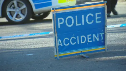 A blue sign with "police accident" in white letters in front of blue and white police tape close to the ground (a tarmac road). The bottom of a police car and two of its wheels are visible in the background as well as a pair of legs walking.