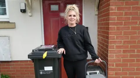 Qays Najm/BBC Kirsty Meads outside of her front door with a black bin to her right and a food caddy in her left hand. She has tied up blonde hair and is wearing a black jumper.