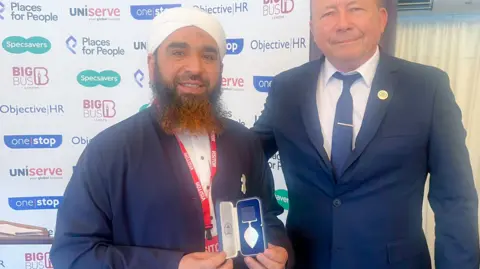 Sameer Hussain Syed Muhammad Faisal Sami holding his award at the ceremony. He has a white turban, a blue top over a white shirt and a long dark beard. He holds a small case with a pointed metal badge attached to a ribbon in it. Next to him is a white man in a blue suit