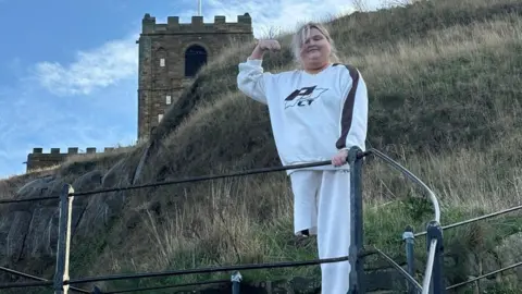 Day One Lucie, a woman with blonde hair and one leg wearing a white tracksuit, stands on the steps outsde Whitby Abbey. 