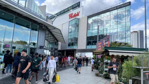 The modern facade and entrance Westfield shopping mall on a sunny day. There are crowds of shoppers walking in and out of the mall. 