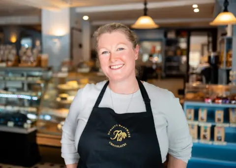Chocolatería Rumsey. Kate Rumsey sonríe en su tienda. Lleva un delantal negro de marca. Detrás de ella se exhibe la variedad de sus productos.