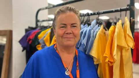 Jasmine DuFraisse/BBC Louise Atherton has brown greying hair and is in a blue top in front of a rail school uniform shirts