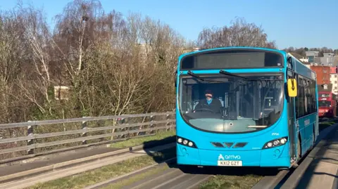 A blue single-decker bus being driven on the guided busway lane, towards the camera. The bus lane is fringed by fencing and trees. 