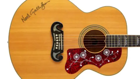 A brown acoustic guitar is pictured, with Noel Gallagher's signature seen on the body of the guitar. Around the sound hole is a detailed red pattern.