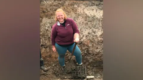 North Pennines National Landscape Rebecca Cadbury-Simmons is standing in a muddy hole in the ground, leaning on a spade and smiling at the camera. She has mid-length ginger hair and is wearing a dark red jumper and jeans.