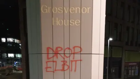 Ben Lafferty A large stone pillar with 'Grosvenor House' engraved in gold and just below 'Drop Elbit' in red spray paint 