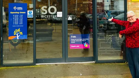 Sobell House A smiling Jay Boydon outside Sobell House shop in Bicester. She is looking at the camera and is pointing towards the entrance. The ground in front is wet.
