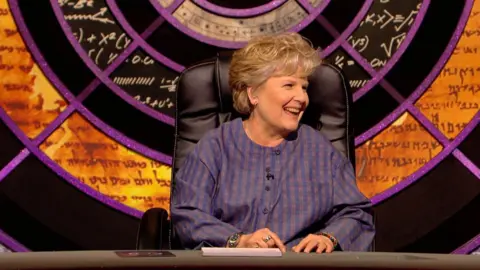 Sandi Toksvig laughs in a blue checked top, on the QI set