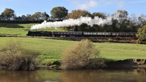 A black steam train pulling three passenger carriages is driving along a track surrounded by fields. There is white steam billowing from its chimney. In the foreground is a river.