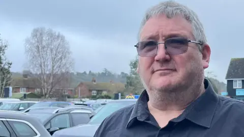 Alan Crossland wearing a dark long‑sleeved shirt is standing in Sandy Town Centre Car Park. Several parked cars are behind him, and trees with bare branches can be seen in the background. Behind the person, across the car park, there is a row of dark‑roofed buildings with shopfronts displaying various signs. 
