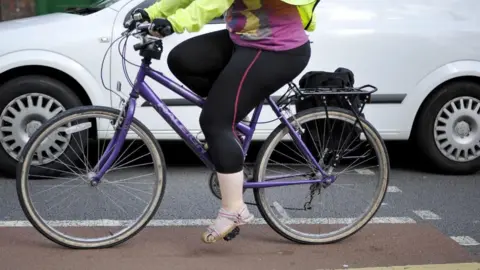 A close-up of the lower half of a woman on a purple bicycle on a road. She is wearing black leggings, sandals, a hi-vis jacket and gloves and there is a white van next to her on the road.