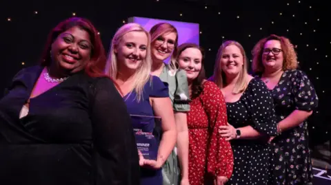 The Mamas Bristol team of six women after their 2024 awards win. They are in front of a black screen with sparkles