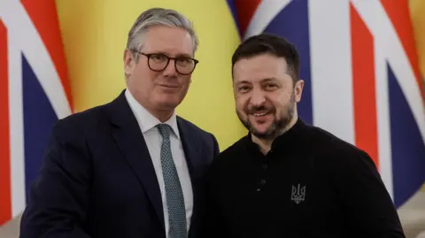 Sir Keir Starmer, with combed-back grey hair and wearing black glasses, a dark blue suit, white shirt and blue patterned tie, stands next to a smiling Volodymyr Zelensky, who is shorter, has short black hair and a beard with a dark shirt.