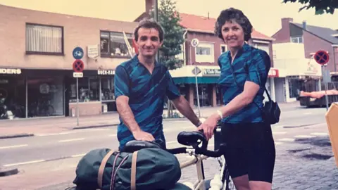Supplied Reg and Pat Marshall in their younger years are on a high street and standing on either side of a bicycle which is piled high with bags. They are both wearing blue cycling tops and black bottoms. Reg, on the left, has short brown hair. Pat has curly, short dark hair. They are both smiling at the camera.