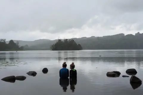 Piers Fulton Two young girls sitting next to each other at the edge of a loch.