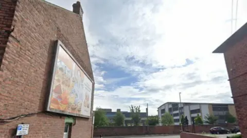 Google The side of a red brick, terraced house with an advertising board on it. The screen has cables coming from behind it, running horizontally across the brickwork. A road can be seen to the side in front of other buildings.