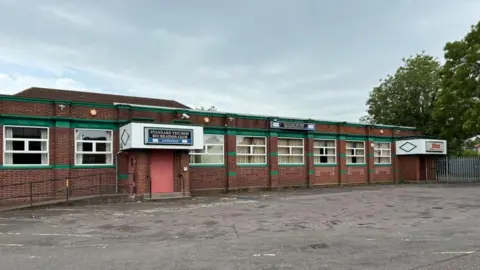 A brick one-storey building with white windows and green painted brick detail with an empty tarmac car park in front. A white porch with Standard Triumph Recreation Club Entrance written above.