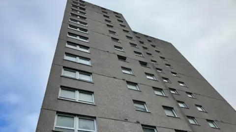 A close up of the tower block, showing the top floors. It is made of grey concrete with white windows. The sky is bright with some clouds.