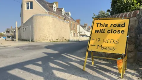 A yellow roadside sign reading “This road will close for 17 weeks from Monday” stands beside a quiet residential street, with stone walls and houses visible in the background on a sunny day.