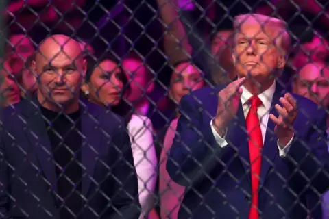 Reuters Dana White and Donald Trump stand next to each other behind a mesh fence with serious expressions. Trump is clapping