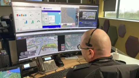 A bald police officer is sat at a desk with four monitors in front of him showing different scenes of an urban area.