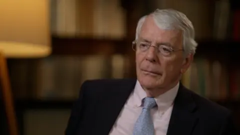 BBC Sir John Major, wearing a dark suit, with a blue tie and white shirt, in a reflective mood