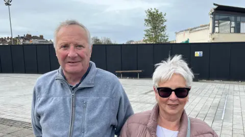 Holly Rattley A man with grey hair wearing a grey hoodie standing shoulder-to-shoulder with a woman with grey hair, sunglasses and a pink coat. Both are standing in front of where the Morrisons supermarket in Swindon used to be - you can see sky space behind them, a large barrier, and the remains of a crumbling building in the background.