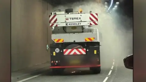 Crane lorry with Terex Kendley company names on the white folded crane seen in the tunnel from the back with grey smoke coming from it and wing mirror of car behind is visible 