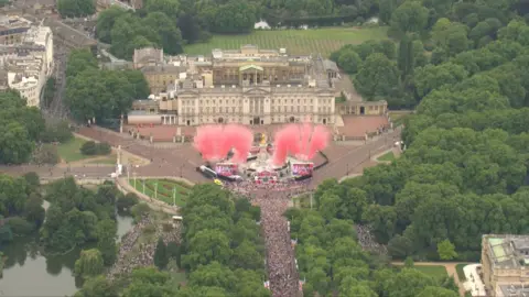 Pink celebratory fireworks emanate from a stage in front of the Queen Victoria Memorial at Buckingham Palace . A large crowd can be seen along the stretch of The Mall. 