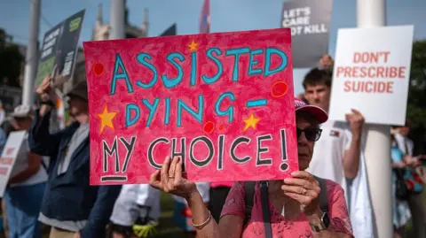 People protest in support of the assisted dying bill at Westminster. A woman is holding a large pink banner saying "assisted dying - my choice" is at the front of the picture. 