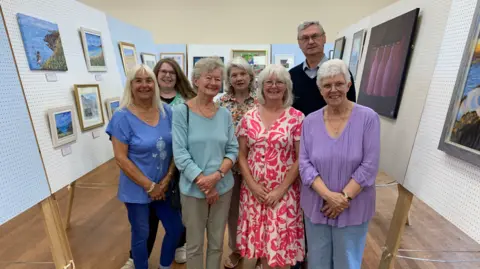 A group of seven members of the Mannin Art group standing in the art exhibition at St Pauls Hall.